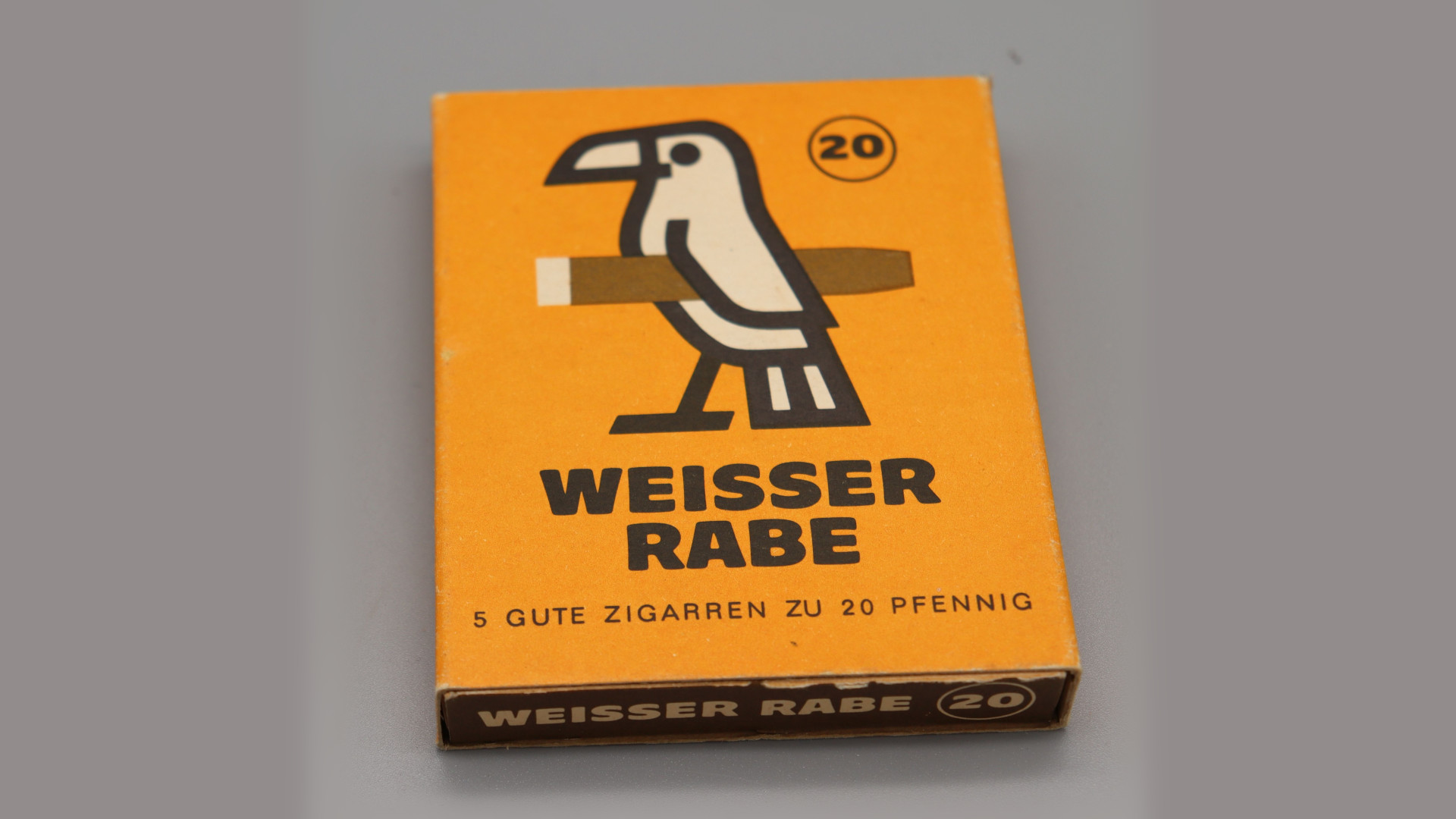 Gelbe Zigarrenschachtel mit der Aufschrift "Weisser Rabe", darüber eine Illustration eines weißen Raben mit Zigarre unter dem Flügel.