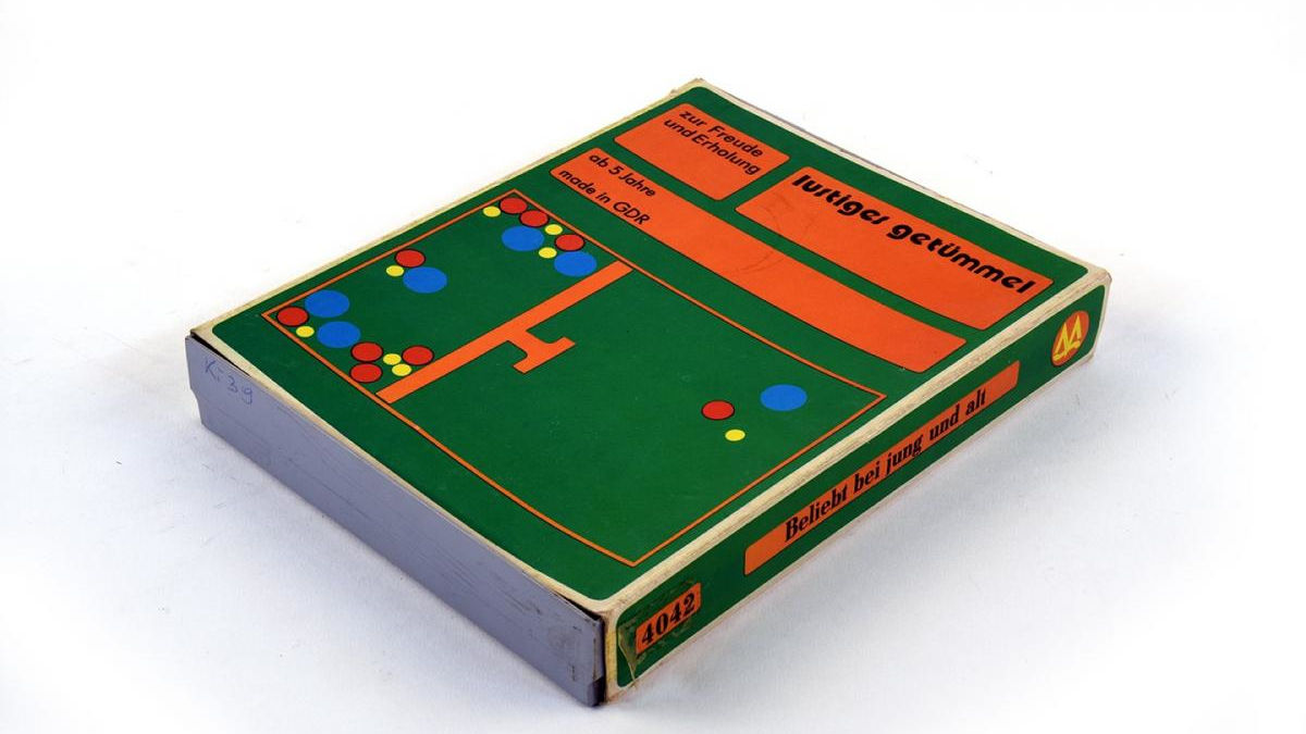 Das Bild zeigt ein altes Tischspiel aus der DDR mit dem Namen „lustiges Getümmel“. Die Verpackung ist in auffälligem Grün und Orange gestaltet und zeigt ein schematisches Spielfeld mit bunten Kreisen in Rot, Blau und Gelb, die vermutlich Spielfiguren darstellen. Auf dem Deckel steht in deutscher Sprache: „zur Freude und Erholung“, „ab 5 Jahre“, und „made in GDR“, was auf die Altersfreigabe ab 5 Jahren und die Herkunft des Spiels aus der DDR hinweist. An der Seite der Schachtel ist der Text „Beliebt bei jung und alt“ aufgedruckt.